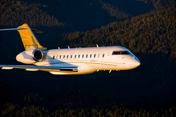 Bombardier Global 5000 in flight over a forested valley at dusk.